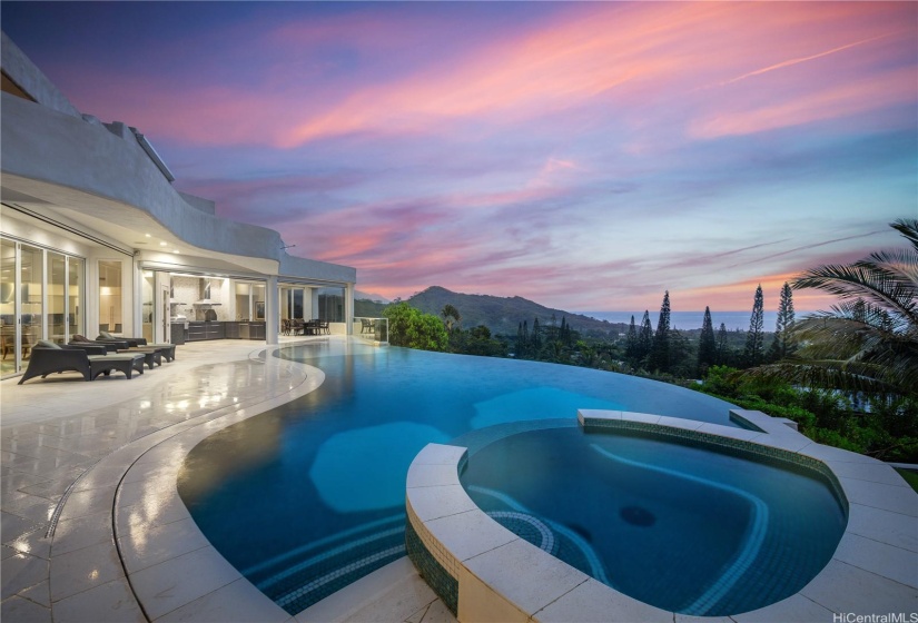 Beautiful infinity pool, jacuzzi, lanai, outdoor kitchen, and outdoor dining area looking out toward the Pacific Ocean at twilight.