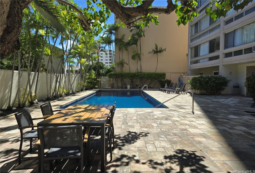 Swimming pool with lounges and tables. Trees provide some shade.