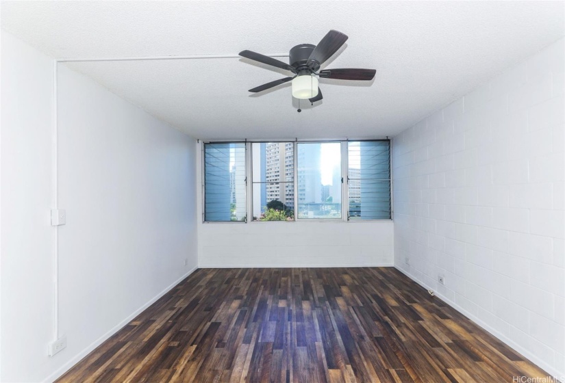 This room features a clean, minimalist design with light-colored walls and a dark wood-style floor, creating a modern yet cozy atmosphere. The ceiling fan adds a functional and aesthetic touch, while the large windows allow for plenty of natural light and offer views of the surrounding buildings, enhancing the room’s open and airy feel.