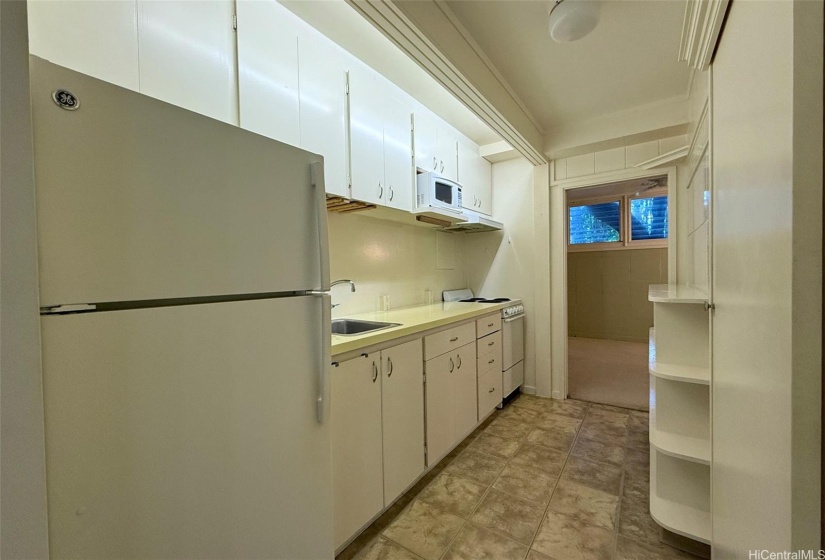 This kitchen has a full-sized refrigerator and extra shelving on the right wall. The view is from the kitchen towards the bedroom.